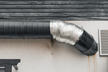 Exterior of an industrial building showing a black ventilation pipe connecting to a silver flexible duct on a textured wall © EdNurg
