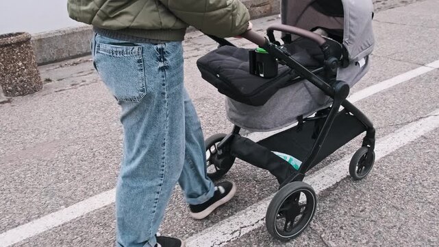 A woman in casual attire is seen from a low-angle shot pushing a baby stroller. The perspective emphasizes the urban sidewalk texture and the motion of the family walk.