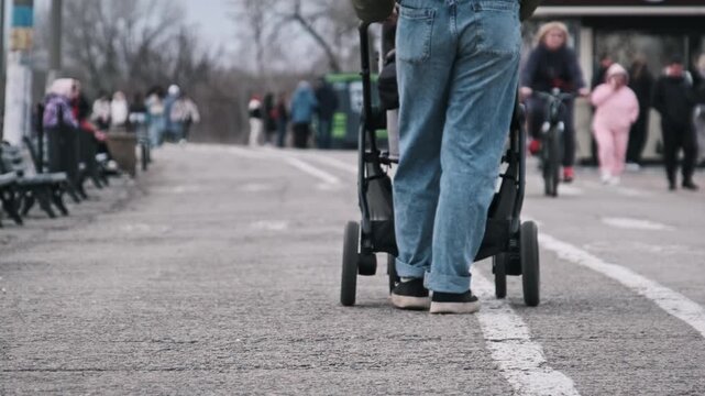 A young mother pushes a stroller along a paved path in a park with other visitors. The low camera angle focuses on the stroller's movement and the mother's walking feet.