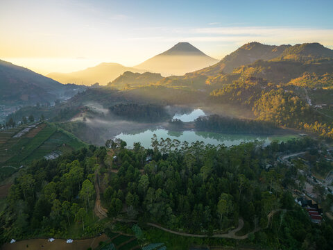 Aerial view of Dieng Park featuring Telaga Warna lake surrounded by lush hills, misty forests, and Mount Sindoro in the background in Kejajar, Central Java, Indonesia.