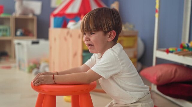 Boy crying with tears on cheek and open mouth, hands gripping an orange stool amid toys and a small tent inside a playroom building; sadness.