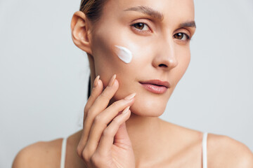 Young woman applies white facial cream on cheek with fingers, has clean skin and natural makeup,...