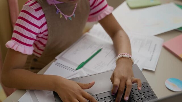 African American child typing on laptop keyboard at desk with homework papers, looking up while studying. Footage perfect for e-learning platforms and online education marketing campaigns