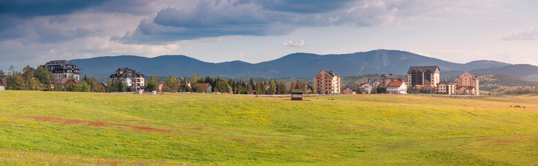 Obraz premium Lush green field in the foreground with a scenic view of traditional and modern buildings in Zlatibor village against mountains, Serbia