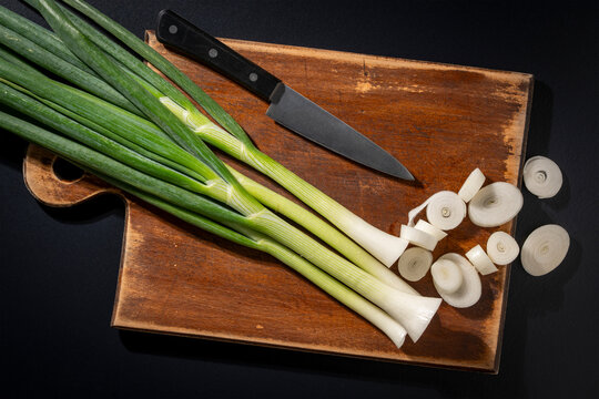 Chopping fresh green onions with kitchen knife on wooden board, closeup. Top view of raw green onion or leek chopped into slices on rustic wooden cutting board next to chef knife on black background