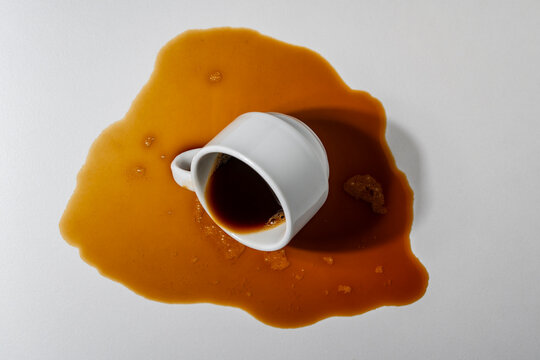 Spilled coffee from overturned white cup on light background, closeup. Top view of a spilled puddle of dark espresso coffee with a fallen white ceramic cup on a white table