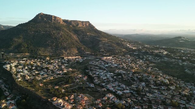 Panoramic aerial view of a sprawling residential area nestled at the base of a majestic mountain range, bathed in the soft glow of dawn. Modern homes blend with lush greenery under a clear sky.