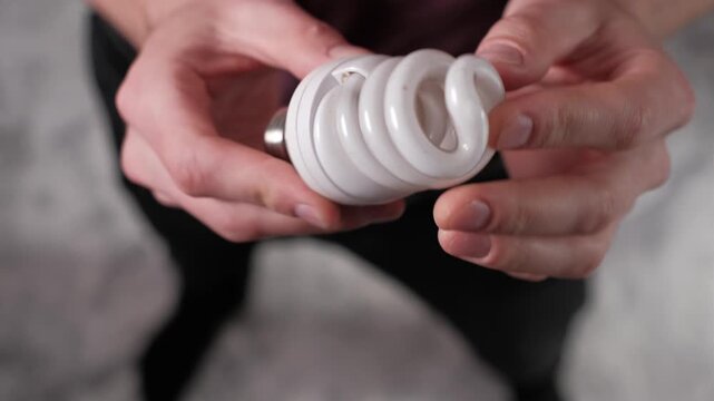 Male hands carefully hold and examine a white spiral compact fluorescent lamp. The twisted tube design and screw base highlight energy-saving light bulb technology.