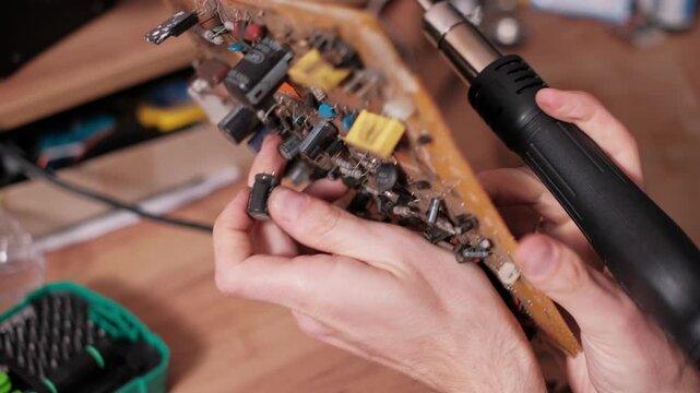 Close-up of hands using a hot air gun station to heat solder joints and remove a capacitor from a circuit board. The focused action depicts precision component replacement and repair.