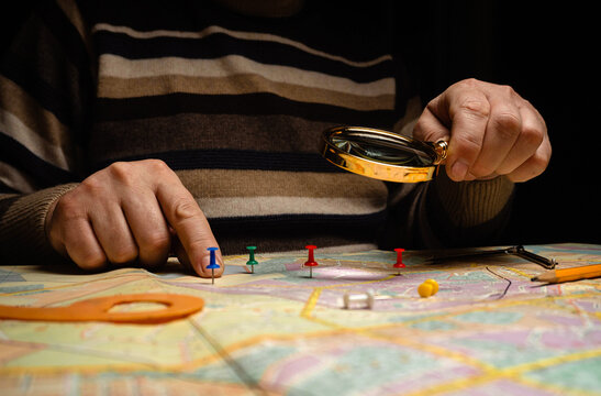 A person uses a magnifying glass to look closely at a map. Colorful push pins mark different locations. The setting is a dim room with a focus on the map and tools