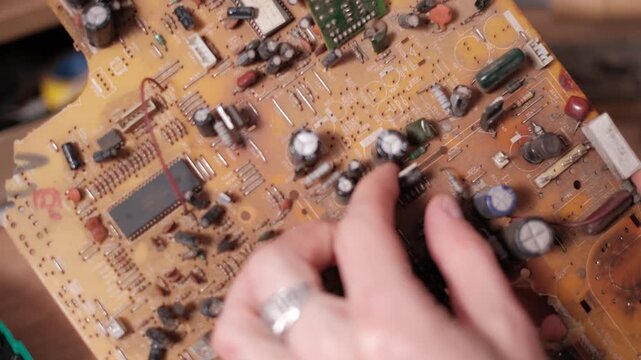 Close-up of hands using a soldering iron to desolder and remove a capacitor from a complex circuit board. The detailed workflow illustrates component replacement and repair precision.