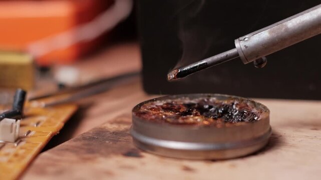 Close-up slow motion view of a hot soldering iron tip smoking as it hovers above a tin of rosin flux. The anticipation of cleaning highlights the preparation phase of precision work.