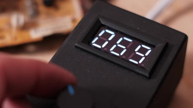 Close-up of a hand turning a knob on a power supply unit to increase the digital LED numbers. The rising value indicates voltage adjustment and precise electronic equipment setup.