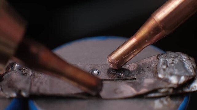 Extreme close-up slow motion of spot welding electrodes joining a metal tab to a battery terminal. Faint smoke appearing post-weld highlights the heat of the industrial connection.