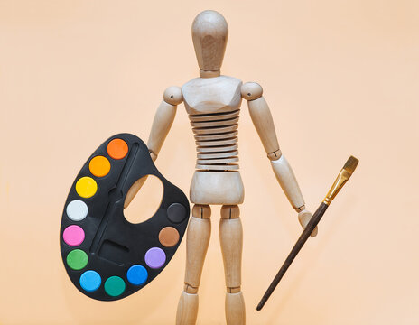 A funny wooden man holds a palette and a brush in his hand on a beige background. Artist and creativity concept.