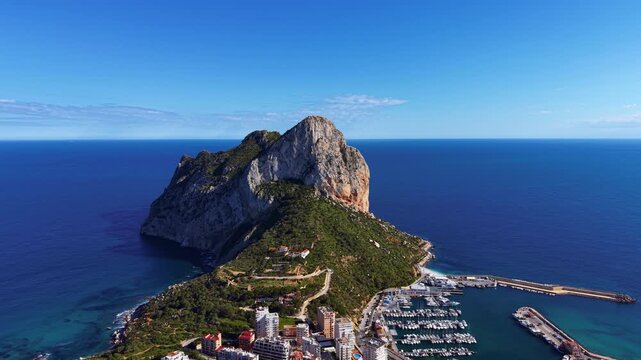 Aerial view of Penon de Ifach natural park rock formation rising above Calpe coastline with marina, residential buildings and deep blue Mediterranean Sea