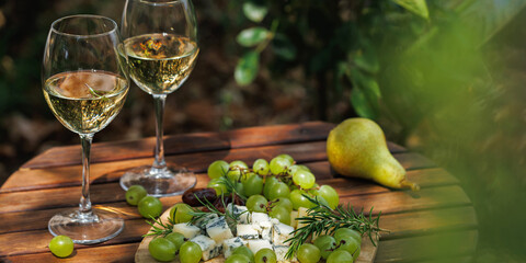Fototapeta premium White wine glasses with cheese board grapes pear and rosemary on rustic table, sunny garden picnic, gourmet food pairing and elegant lifestyle