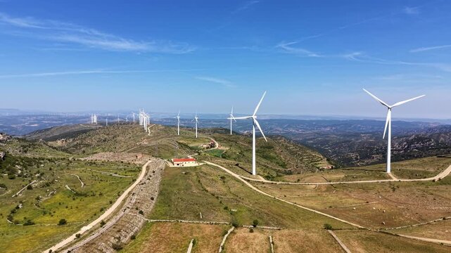 Aerial view of multiple wind turbines spread across green rolling hills with winding dirt roads, small farm buildings, and expansive valley landscape under a clear blue sky