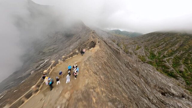 Cinematic FPV drone flight along the rim trail of Mount Bromo crater before diving into the steaming volcanic crater. Tourists hike the narrow ridge in East Java, Indonesia.