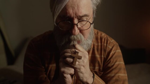 Chest up of senior Caucasian man holding wooden cross, praying alone at home. Clip is suitable for religious organizations, mental health support, and senior care marketing campaigns