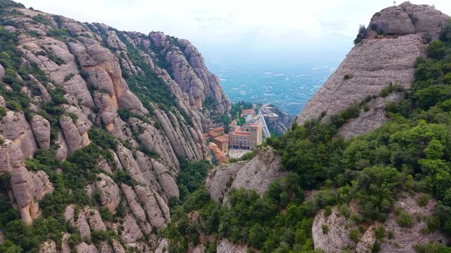 High angle view down the valley over Montserrat monastery complex flanked by vertical conglomerate rock faces, with the misty Catalan plain stretching into the distance