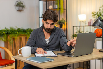 Obraz premium Indian man at home compares paper files with laptop spreadsheet writes notes stays focused. Arabian guy at desk checks rows and totals reviews values does careful review and confirms match calm mood.