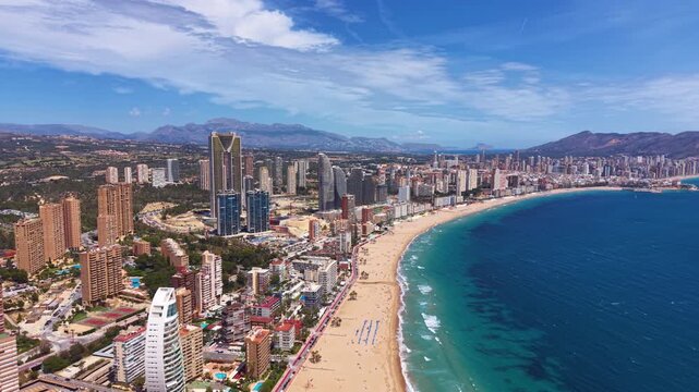 Panoramic aerial view of Benidorm, Spain, showcasing its iconic skyscrapers, golden sandy Levante beach, turquoise Mediterranean water, and mountain backdrop on a sunny day