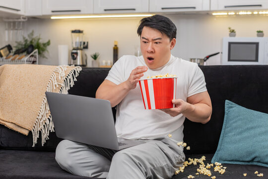 Asian man at home streams tense film on laptop flinches at jump scare drops popcorn steadies breath. Chinese guy on sofa resumes watch picks pieces of snacks holds backet, keeps focus for tense scene