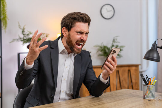 Middle-aged businessman at home office spends money on online lottery casino smartphone app groans as luck fails. Freelancer guy slams fist hard exhales heavy drops head on hands after gambling loss