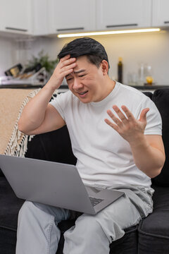 Asian man at home sees game loss on laptop touches forehead sighs and hits retry with focus. Chinese guy on sofa plans rematch sets target adjusts settings breathes and gets ready calm again steady