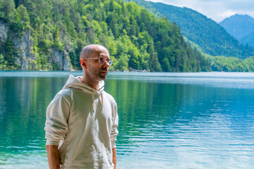 Fototapeta premium Bald young man wearing glasses, white hoodie, plaid trousers, Travel and tourism stylish clothing choices, pebble beach, turquoise lake, mountains, forest, summer vacation, travel, fashion portrait