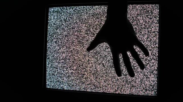A dark silhouette of a hand touches a flickering screen filled with chaotic television static noise. Concept of supernatural mystery, tech glitch, paranormal activity, and retro horror.