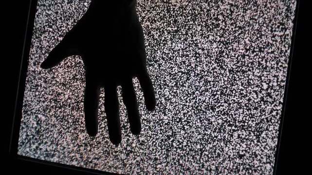A dark silhouette of a human hand touches a glowing television screen filled with heavy static noise. Concept of paranormal activity, lost signal transmission, and retro technology glitches.