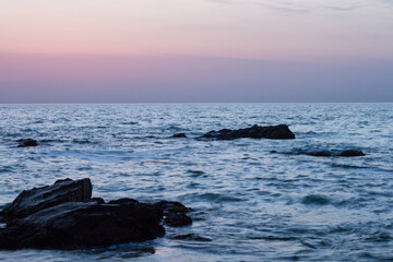 Obraz premium Serene ocean waves gently lap against dark rocks at twilight with a soft pink and purple sky