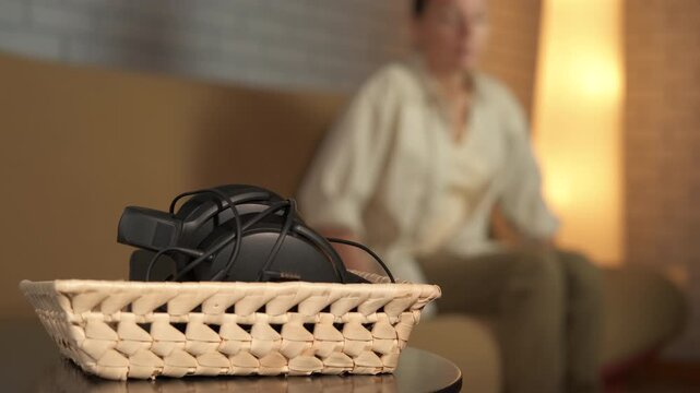 Woman grabbing headphones from basket to listen music. Relaxed woman sitting on a sofa gets up and takes her over-ear headphones from a basket on a side table, ready to enjoy some audio entertainment