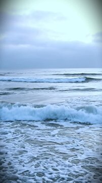 Waves crash onto the sandy beach shoreline under a cloudy sky with a gloomy atmosphere and ocean water.