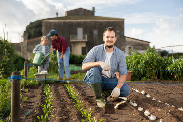 Obraz premium Male farmer plants garlic in the garden with his family on the background. Farmer has planted and grows organic seeds.