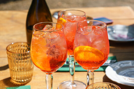 Three glasses of refreshing orange spritz cocktails with ice and citrus slices served outdoors on green sunny patio. Summer relaxation, outdoor dining, leisure, travel and social gathering concept.