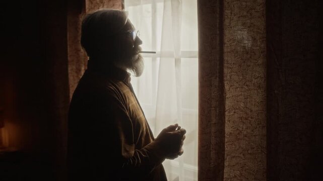 Side view medium shot of senior Caucasian man standing by window, lighting cigarette and holding mug. Perfect for commercial campaigns addressing senior loneliness, mental health, or aging habits