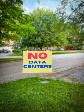 Vertical, close-up shot of a "NO DATA CENTERS" community protest sign placed in a grassy front yard, with a blurred suburban neighborhood street in the background.