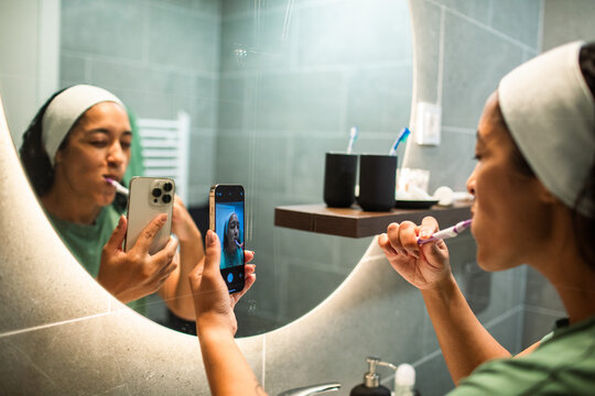 Young woman brushing teeth while recording a mirror selfie in home bathroom