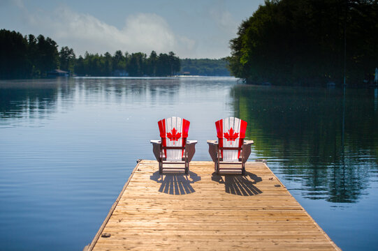 Two Adirondack chairs painted with the Canadian flag sit on a wooden dock overlooking a calm Muskoka lake, framed by forested shores under a soft blue summer sky. Canada Day cottage country.