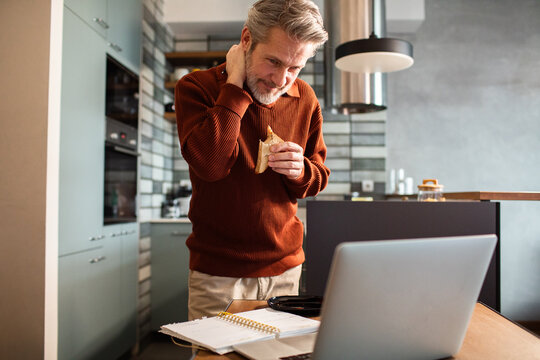 Middle-aged man rubbing neck while working on laptop in home kitchen