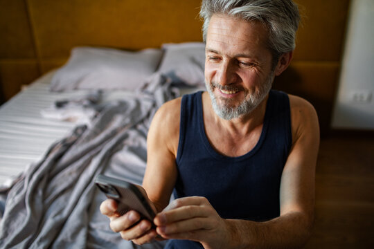 Smiling middle-aged man using smartphone in home bedroom