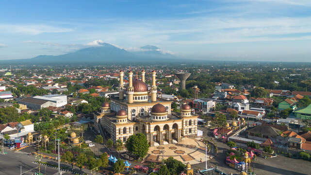 Aerial view of the Great Mosque of Klaten with its prominent domes surrounded by the urban landscape and a distant mountain under a clear blue sky in Indonesia.