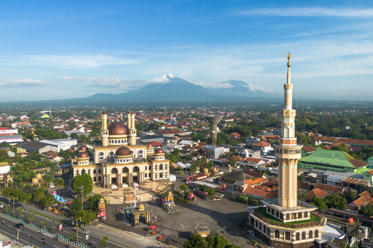 Aerial view of the Great Mosque of Klaten with its dome and minaret, surrounded by the urban landscape, with a distant mountain under a clear blue sky in Indonesia.