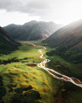 Aerial view of the winding river flowing through the lush green Turgen Valley surrounded by majestic mountains under a soft cloudy sky in Turgen, Kyrgyzstan.