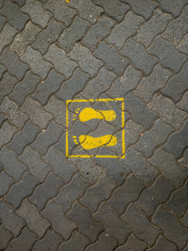 Yellow footprint sign painted on gray paving blocks, indicating the designated standing position for people waiting in a queue or maintaining physical distance