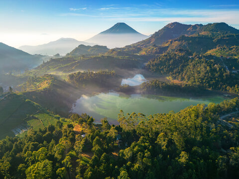Aerial view of Dieng Park featuring the volcanic lakes surrounded by lush green hills and mountains under a clear sky in Kejajar, Central Java, Indonesia.