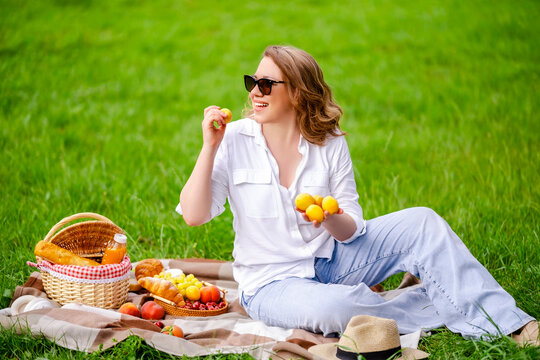 Cheerful woman with fresh apricot on picnic blanket in summer park. Fruits, sunglasses, wicker basket, healthy snack and summer nutrition concept.